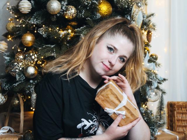 Jessika Daniels mit blonden Haaren im Pferdeschwanz, die ein verpacktes Geschenk vor einem geschmückten Weihnachtsbaum hält, trägt ein schwarzes Hemd und lächelt sanft..
