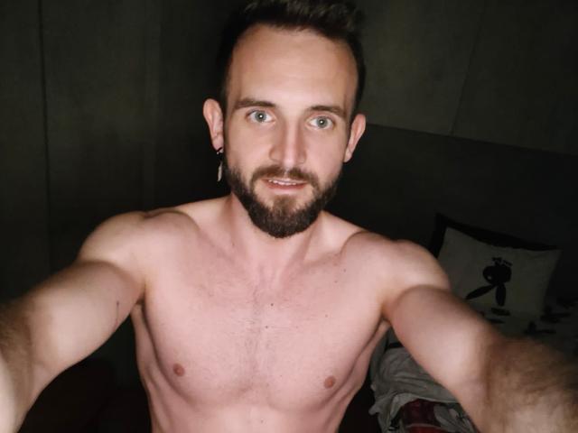 TonyHotX, ohne Hemd, mit muskulösem Körperbau, kurzen braunen Haaren und Bart, hat blaue Augen und kleine Ohrringe. Er hält eine Kamera für ein Selfie, wobei er seine Brust und Arme zeigt..