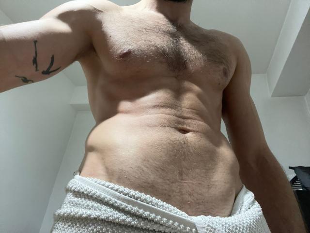 Il torso nudo e muscoloso di TonyHotX, con il petto peloso e un piccolo tatuaggio sul braccio sinistro, indossa un asciugamano bianco intorno alla vita, mostrando un pene leggermente visibile, flaccido e non circonciso..