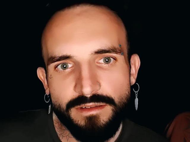 TonyHotX tiene la cabeza calva, ojos verdes, un piercing en la nariz, aretes, barba y un tatuaje en el cuello. Su rostro está ligeramente inclinado y tiene una expresión seria..