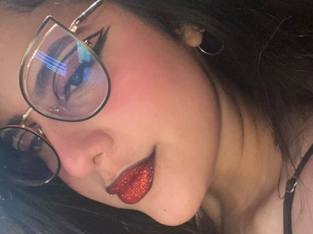 ChloePrie with red glitter lips, black eyeliner, and glasses, showing her tongue. She has dark hair, hoop earrings, and a slight blush on her cheek.