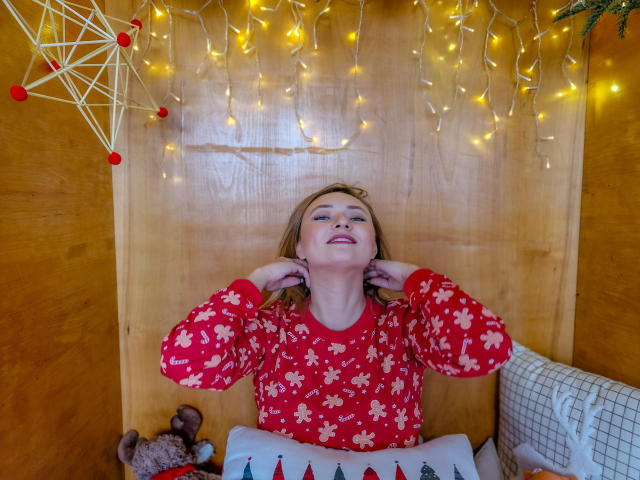 SallyVerda with blonde hair, red floral pajamas, hands behind head, smiling, wooden wall, string lights, stuffed animal, pillow with trees, and fairy lights above.