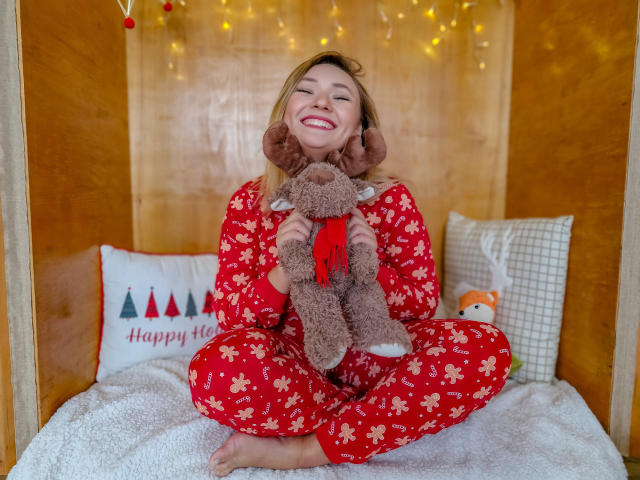 SallyVerda in red floral pajamas, smiling, holding a brown stuffed reindeer with a red scarf, sitting cross-legged on a white bed with wooden walls and fairy lights.