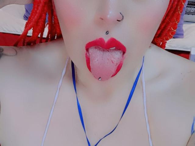 IsabellaRoshe with red braids, nose ring, tongue piercing, and red lipstick, tongue out, blue necklace, pale skin, and visible cleavage. Her tongue is covered in saliva.