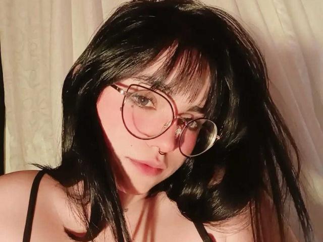 LovelyLilith with black hair, round glasses, nose ring, pink skin, and a serious expression, showing her tits in a black spaghetti-strap top.