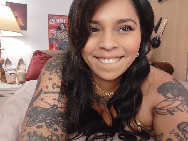 Chili, smiling with her tanned, tattooed shoulders and large breasts visible, wearing headphones, leaning on a bed.