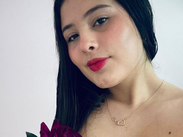 Kathy Stone, met lang zwart haar, rode lippenstift en een gouden hartketting, houdt een paarse roos vast en kijkt iets opzij met een subtiele glimlach..