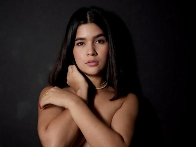 ScarlletZhen with dark hair, crossing arms over bare tits, looking at camera, wearing a gold necklace, dark background, red nail polish, gold ring, soft light.