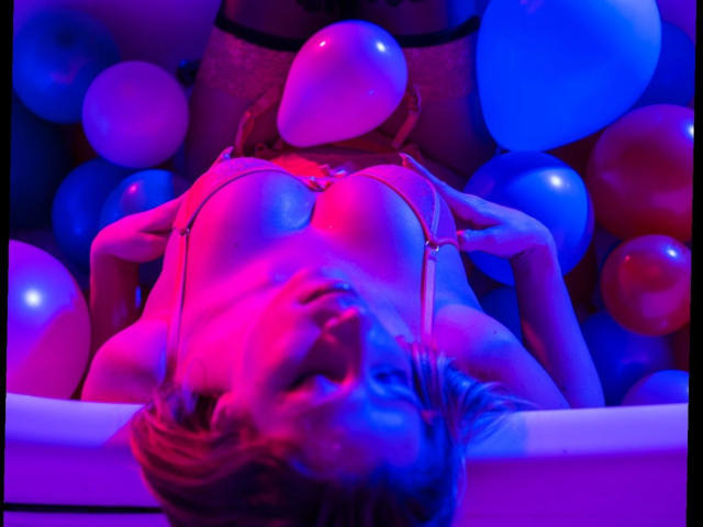 Livie-hot with big tits, lying in a tub of colorful balloons, wearing a red bra, illuminated by pink and blue lights, looking up with her head tilted back.