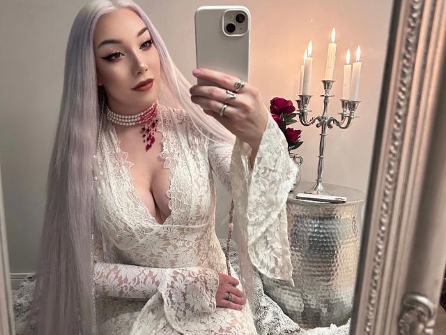 Lady-Khaleesi with long, white hair, wearing a lace, low-cut, white dress, showing her tits, taking a selfie, holding a phone, with a silver candelabrum and red roses.