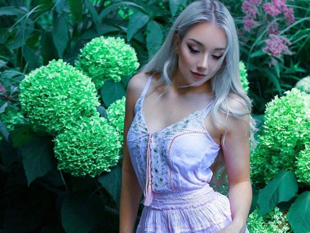 Lady-Khaleesi with blonde hair, wearing a lilac lace-up dress, standing among green hydrangeas, looking down with a subtle smile, showing her small tits and slim ass.