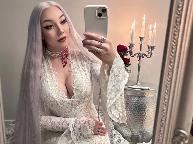 Lady-Khaleesi with long, white hair, wearing a lace, deep-cleavage white dress, holding a phone, showing her tits, and a red necklace, with a candleholder and roses.