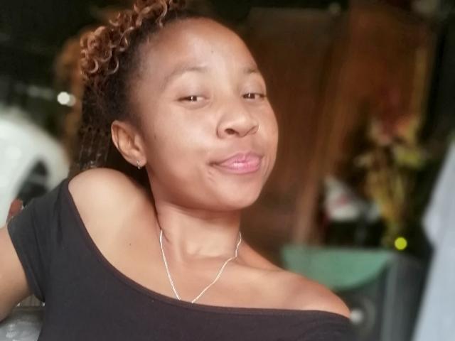 Edrwina69 has medium brown skin, curly brown hair, and is wearing a black off-shoulder top with a silver necklace, pouting her pink lips. Her tits are small.