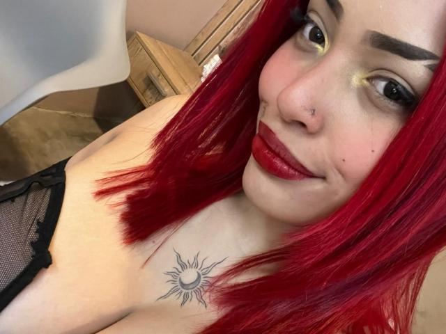 StrawberryX with red hair, nose ring, and sun tattoo on chest, wearing black mesh top, red lipstick, and yellow eyeshadow, smiling at camera. Boobs visible.