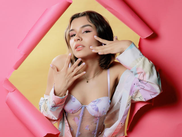 LenaBranch, con las manos en el rostro, vistiendo una blusa de encaje rosa con estampado floral que deja ver sus pequeños senos, y una bata floral en tonos pastel, sobre un fondo rosa y amarillo..
