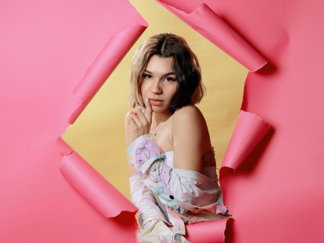 LenaBranch with blonde and brown hair, wearing a floral off-shoulder top, biting her finger, pink and yellow paper strips framing her, looking sexy and confident.