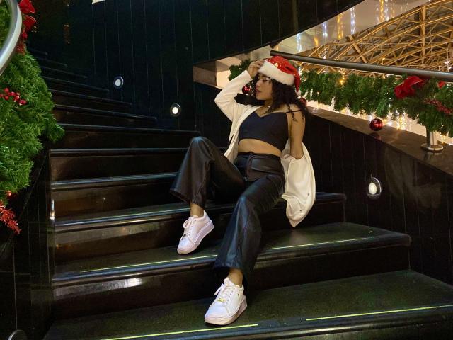 SophieSonyas sits on black stairs wearing a red Santa hat, white jacket, black crop top, leather pants, and pink sneakers, covering her eyes with her hand.