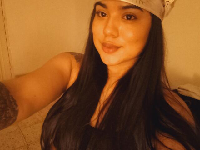 SteffanyTaylor with long black hair, wearing a white cap, has full tits, tattoo on her right arm, and a slight smile, in a warm, yellow-lit room.