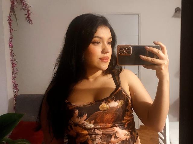 LauraRichis with long black hair, wearing a brown floral tank top, holding a phone, taking a mirror selfie, showing her tits, slightly pouty lips, and warm skin.