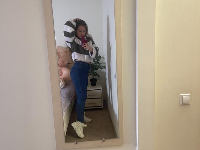 Dooris with striped sweater, blue jeans, yellow socks, holding phone, mirror selfie, standing by bed with pink teddy, hand in hair, room with white walls and wooden floor.
