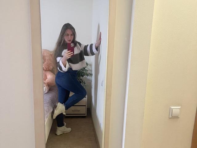 Doris se tient devant un miroir, vêtue d'un pull rayé, d'un jean bleu et de baskets blanches, tenant un téléphone rouge, une main appuyée contre le mur, dévoilant ainsi ses fesses..