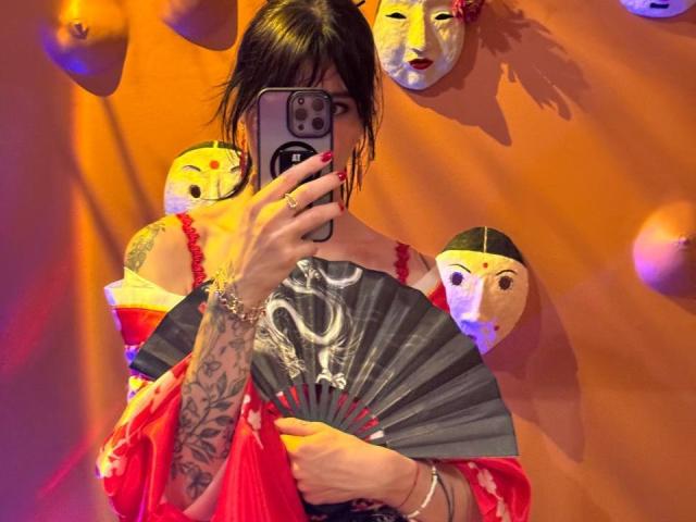 Tsukerberg mit Tattoos, der einen Fächer hält, in einem roten und pinken Kleid, Spiegel-Selfie, orangefarbene Wand mit Maskenköpfen, dunkles Haar, Telefon, das das Gesicht verdeckt, rote Nägel, Armbänder..