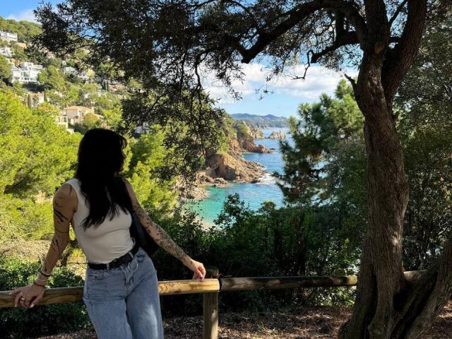 Tsukerberg with black hair, white tank top, blue jeans, and tattoos, leans on wooden railing, facing ocean view, standing under tree, sunny, green foliage, clear blue water, rocky coast.