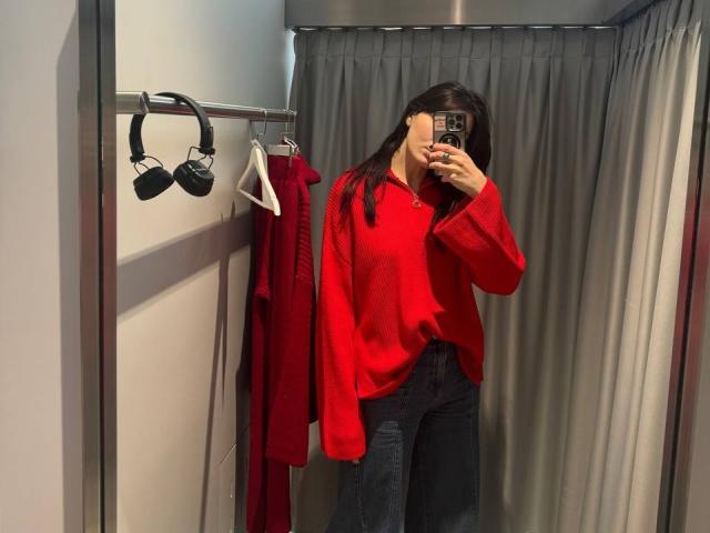 Tsukerberg with long dark hair, wearing a red sweater and black pants, taking a mirror selfie in a changing room. Red dress and black headphones on a rack.