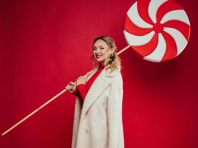 SallyVerda with red lipstick, blonde hair, white coat, red sweater, holding giant candy cane swirl, red background, smiling, standing, wood candy cane, tits slightly visible under coat.