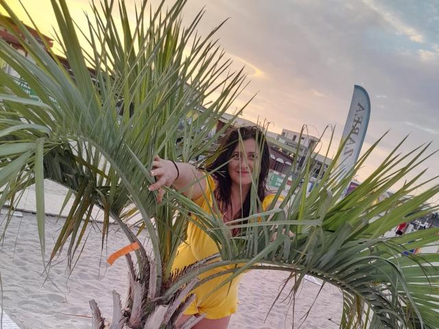 BlissSophia with long brown hair, yellow top, and red lipstick, leaning through green palm fronds, smiling, on sandy beach, sunset sky, holding palm leaves, orange ribbon on trunk.
