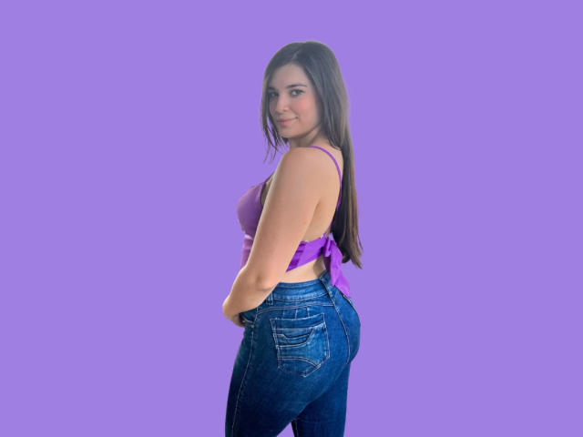 Katelyn Allen, met lang bruin haar, een paars topje met losse mouwen dat haar grote borsten laat zien, en blauwe jeans, staat zijwaarts en kijkt terug met een lichte glimlach. Paarse achtergrond..