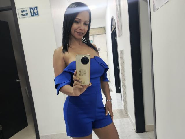 EvelinFoster with black hair, blue off-shoulder dress, holding phone in hallway, smiling, showing her tits and ass in mirror selfie, white walls, beige tiles, and black doors.