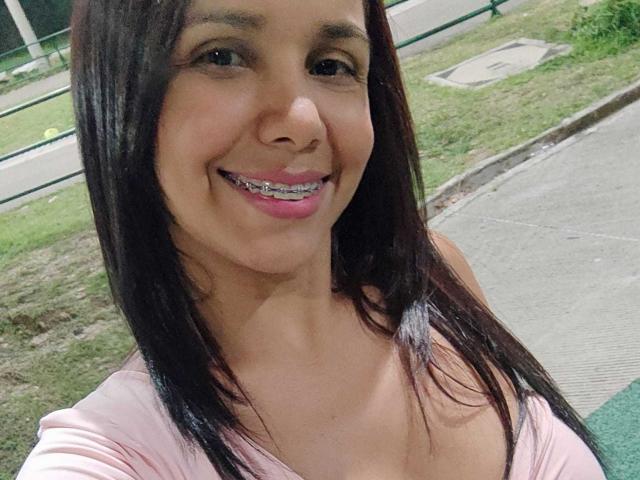 EvelinFoster with straight black hair, pink lipstick, and metal braces on her teeth, smiling at the camera in a pink top, showing her tits and cute face.