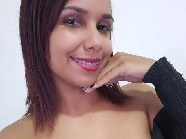 EvelinFoster has dark brown hair, pink lipstick, and is smiling with her hand near her chin. She's wearing a black, ribbed, off-shoulder top. Her teeth are metal.