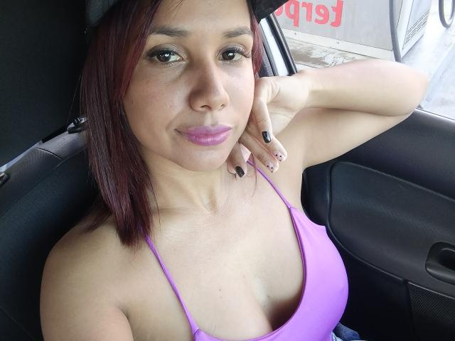EvelinFoster with red-brown hair, purple lipstick, black nails, and purple tank top showing her small tits, sitting in a car, arm behind head, looking at camera.