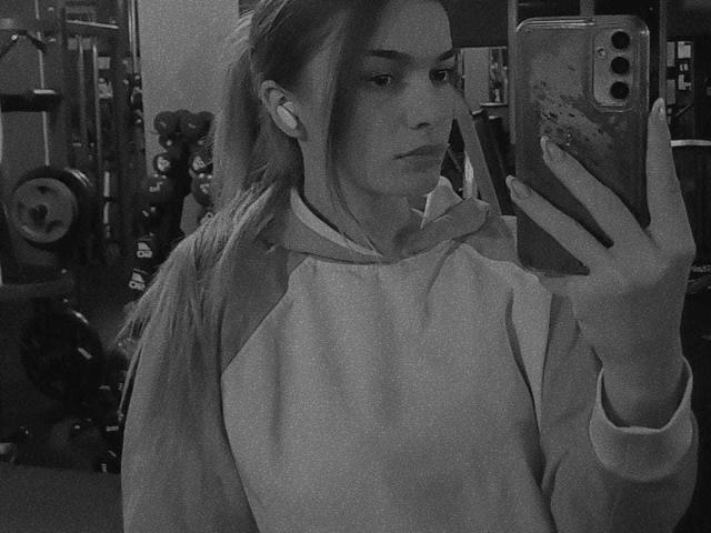 IngridI with long hair in a ponytail, wearing a raggedy hoodie, holding a phone with a dirty case, looking in a mirror, wearing earbuds, in a gym.