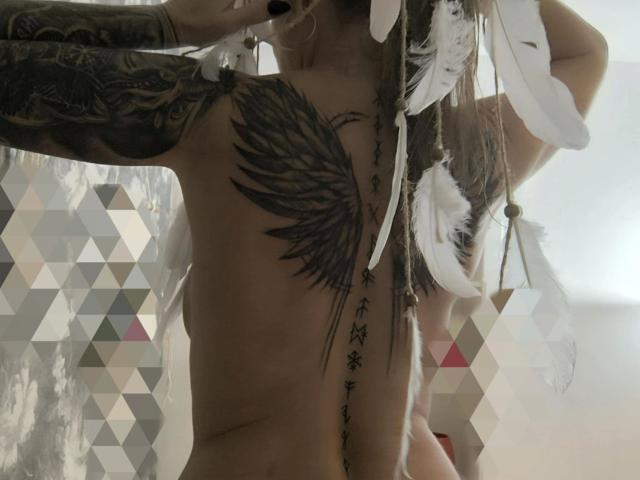 Nude KatyStar-hot with tattoos, white feathers in hair, black nipple plugs, and a large wing tattoo covering her tits. Her arm tattoo is detailed, with Chinese characters on her belly.