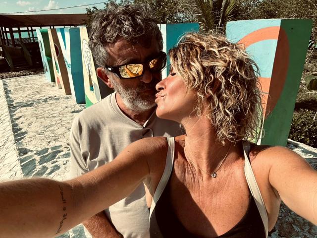 AngeelCouple selfie: older dude with gray beard and sunglasses, kissing AngeelCouple's sun-kissed, curly blonde head, tan top, necklace, tan arms, outdoor, colorful wall in background.