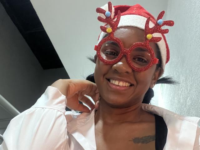 TiffanySammy with dark skin, smiling, wearing red glitter reindeer glasses, Santa hat, white shirt, black strap, and visible tattoo, touching cheek with hand.