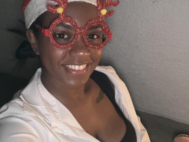 TiffanySammy with dark skin, smiling, wearing red glitter reindeer glasses, white hat, white jacket, black top, showing small tits, and a tattoo on her right shoulder.