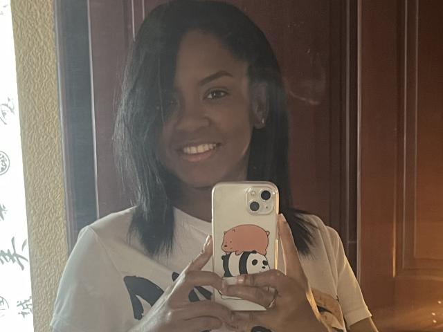 TiffanySammy with dark skin and black hair, smiling, holding a phone with a pink bear case, wearing a white shirt, taking a mirror selfie.