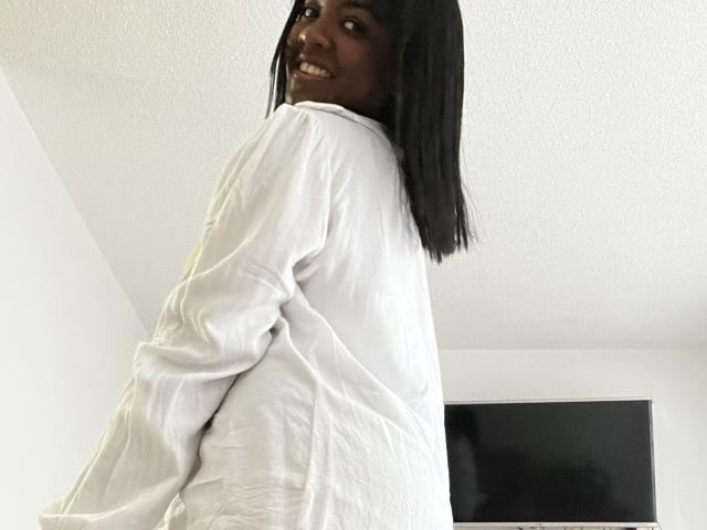TiffanySammy, mit dunkler Haut, glattem schwarzem Haar, lächelnd, trägt ein lockeres weißes Hemd, steht in einem weißen Raum, blickt über ihre Schulter und zeigt ihr Gesäß..