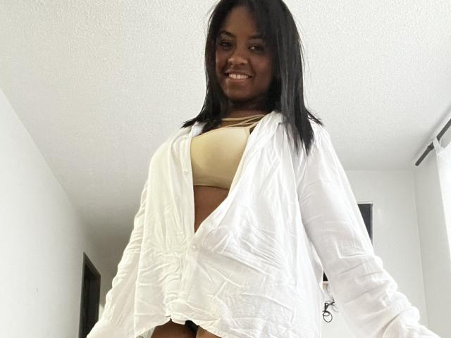 TiffanySammy, com pele escura, cabelo preto liso, sorrindo, vestindo uma camisa branca de botões por cima de um top esportivo bege, mostrando sua barriga plana e seios pequenos e arredondados..