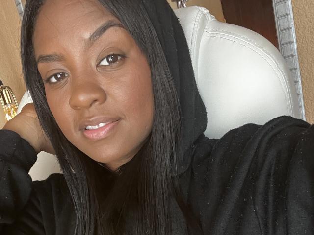 TiffanySammy has dark brown skin, straight black hair, and brown eyes. She's wearing a black hoodie with her head tilted, smiling slightly with a relaxed, confident look.