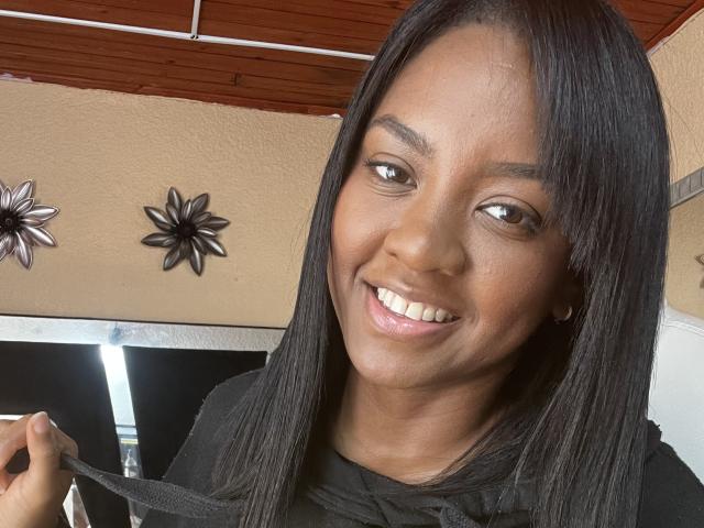 TiffanySammy with smooth dark skin, straight black hair, and a big smile, holding a black shirt's edge, showing white teeth, and brown eyes, wearing small gold earrings.