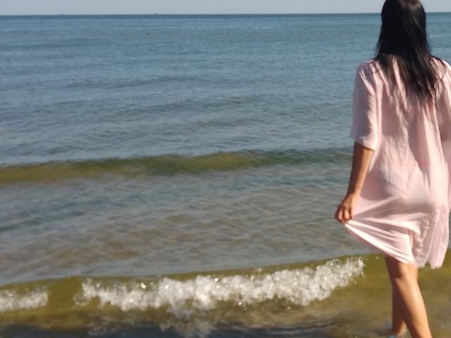 MirandaHott69 with long black hair, wearing a pink dress, stands in shallow water, lifting the dress with her right hand, showing her ass. Ocean and small waves in background.