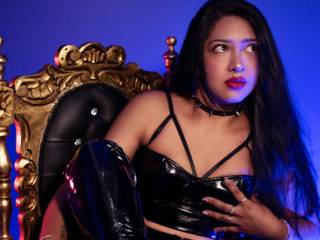 Melanie Morel, con cabello negro largo, vestida con un top corto negro brillante, un collar y un brazalete de cuero negro, sentada en una silla de oro ornamentada, mirando hacia un lado y con lápiz labial rojo..