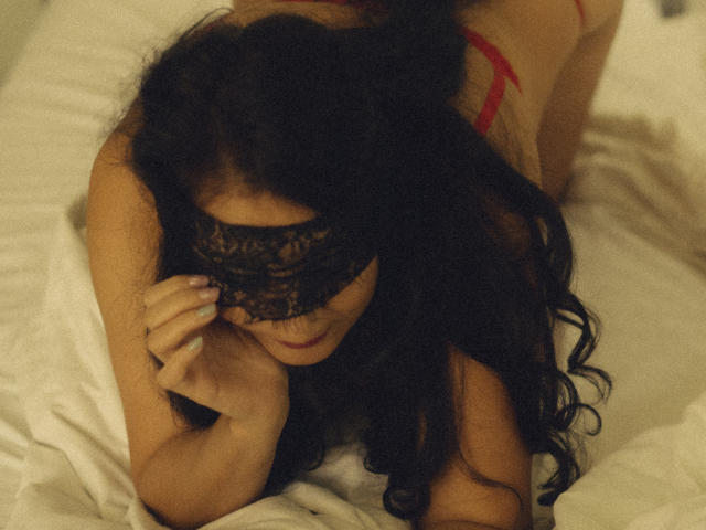 KarinaCecillia with long black hair, wearing a black lace mask, red bra, and nothing else, on a bed, covering her eyes with her hand, in a sensual pose.