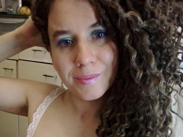 SelenaXCute with curly brown hair, blue eyeshadow, pink glossy lips, white lace top, smiling, hand in hair, visible armpit, slight blush, cute, attractive, soft lighting.
