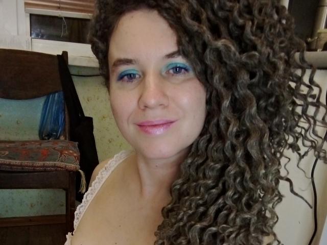 SelenaXCute has blue eye makeup, curly brown hair, pink lipstick, and a slight smile, wearing a white lace top, showing her tits and ass in the photo.